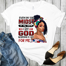 Load image into Gallery viewer, White t-shirt with a young African American black woman wearing a head band and a cross. With the words even in the midst of the storm, I see God working it out for me. Also in the picture is black shoes and blue jeans on the bottom sides.
